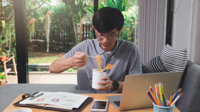 Asian man eating instant noodles while working on laptop at his home office.